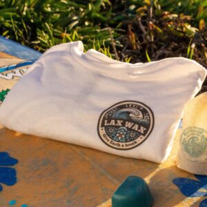 Lax Wax eco-friendly surf wax t-shirt folded on a surfboard at the beach with reusable wax bags and wax bar in sunlight