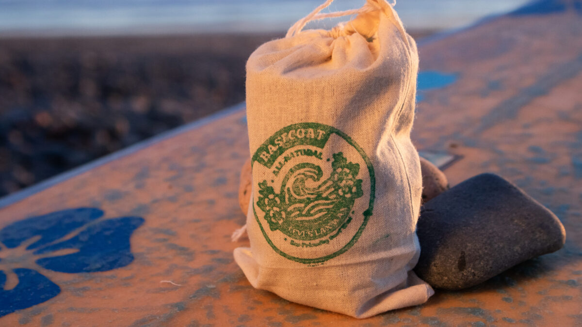 A canvas bag with a Lax Wax logo sits on a weathered surfboard beside a smooth stone, bathed in warm golden sunset light with a blurred beach background.