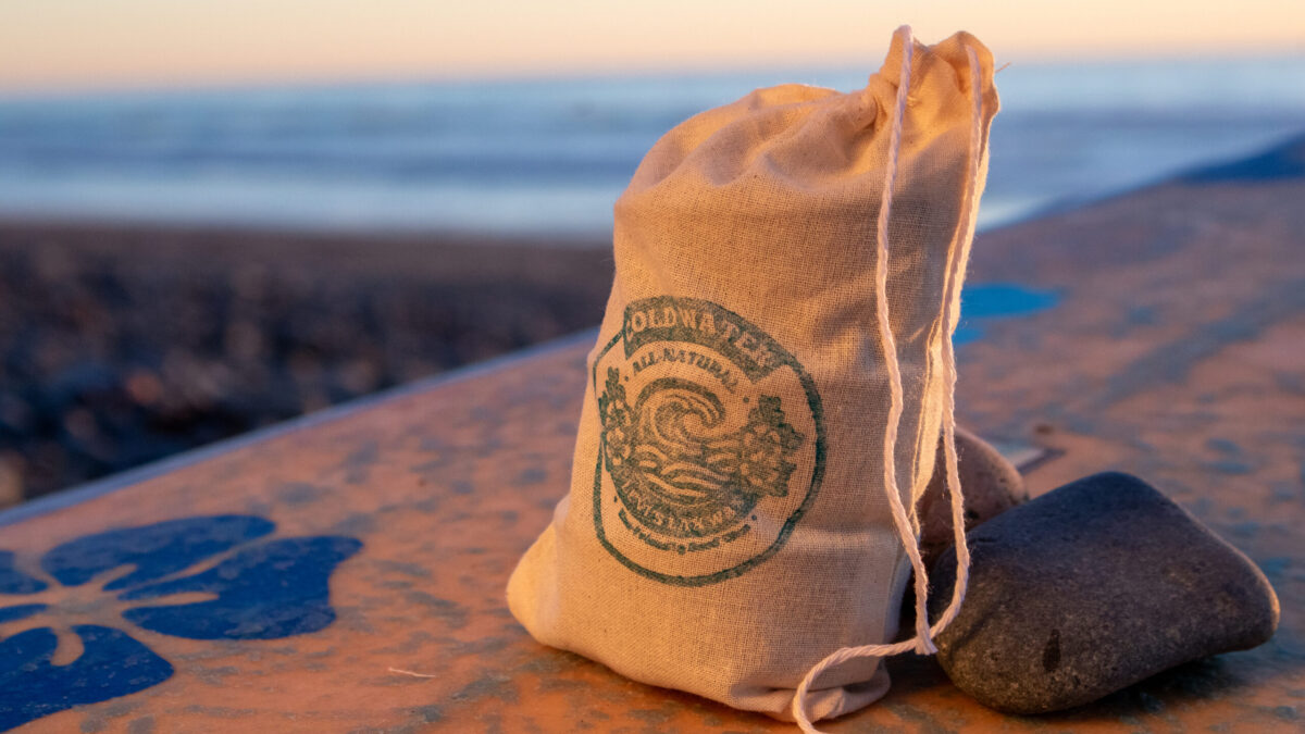 A canvas bag with blue Lax Wax cold water logo rests against a smooth stone on a weathered surfboard at sunset, glowing in warm orange light with a soft-focus ocean and beach in the background.