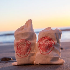 Two canvas bags with red Lax Wax warm water logos stand on wet sand at the shoreline during sunset, with soft ocean waves and a glowing yellow-orange sky in the background.
