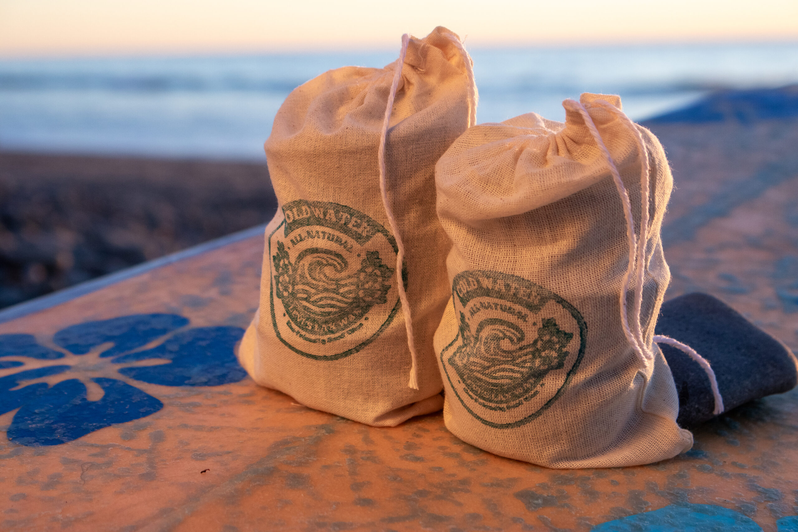 Two canvas bags with Lax Wax logos sit on a surfboard at golden hour, with soft ocean waves and warm sunset light in the background.