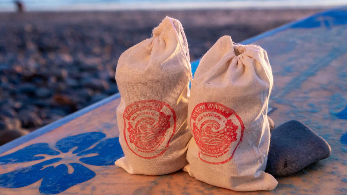 Two canvas bags with red Lax Wax warm water logos rest on a weathered surfboard with blue graphics, with a rocky beach and overcast sky in the background.