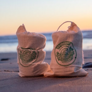 Two tied canvas Lax Wax bags sit on a beach at sunset, with ocean waves and soft orange-pink sky in the background.