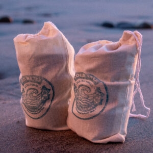 Two tied canvas bags with Lax Wax cold water logos stand on wet sand at the water's edge, with gentle waves and scattered rocks in the soft blue-hour light.