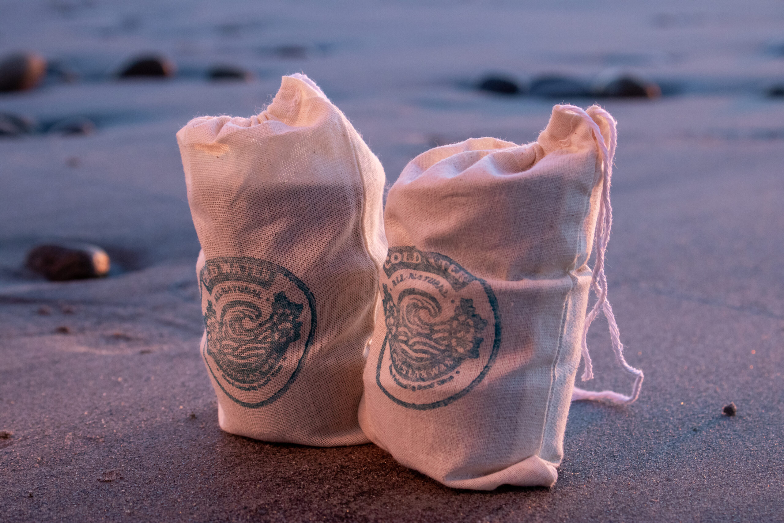 Two tied canvas bags with Lax Wax cold water logos stand on wet sand at the water's edge, with gentle waves and scattered rocks in the soft blue-hour light.