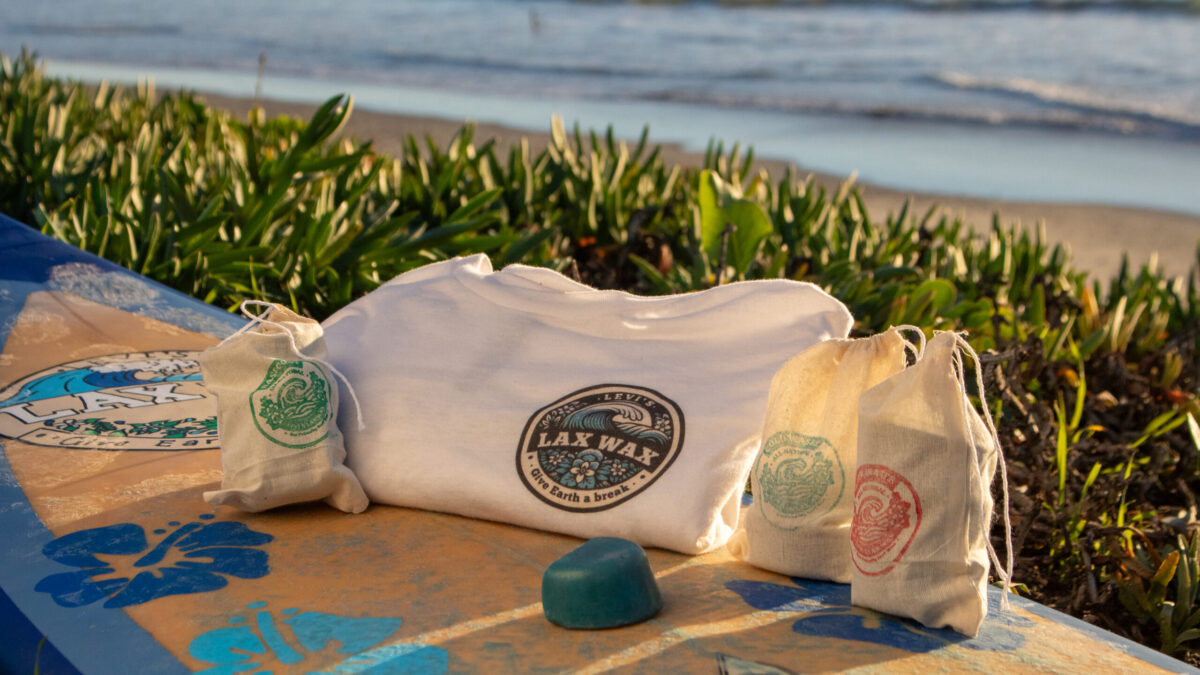 Lax Wax product lineup on a weathered surfboard at the beach during golden hour: two turquoise wax bars in front, a white t-shirt with Lax Wax logo in the center, and canvas bags with green, red and blue Lax Wax logos on either side, with ocean and coastal plants in the background.