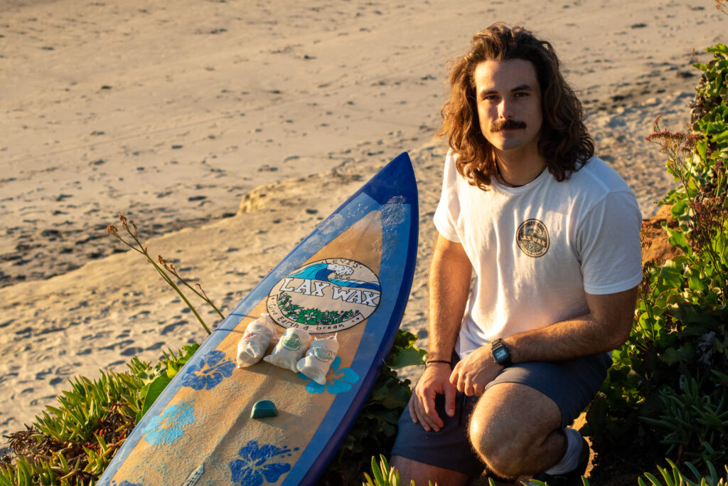 A surfer with shoulder-length wavy hair wearing a white Lax Wax t-shirt sits on sandy beach next to a blue Lax Wax surfboard displaying Lax Wax products and canvas bags, with coastal vegetation framing the scene in warm golden light.