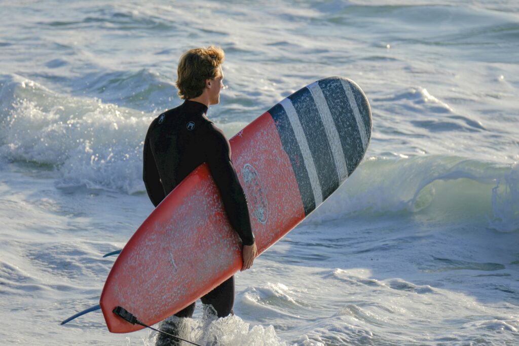 Long Board - Surf Wax A surfer in a black wetsuit standing in shallow ocean water, holding a red longboard with dark blue and white horizontal stripes on the deck. The surfer is viewed from behind, watching incoming waves. Water droplets are visible on the waxed surface of the surfboard, and white foam from breaking waves surrounds the surfer's legs - Surf Wax