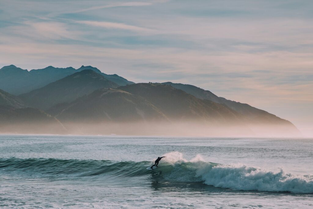 Surfer riding a breaking ocean wave with dramatic misty mountain ranges and a soft pastel sky stretching across the horizon in the background - Eco Friendly Surf Wax
