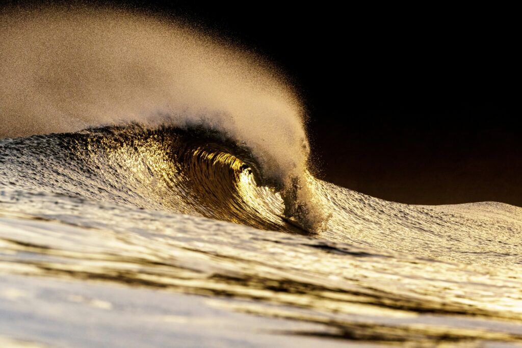 Wave Barrel - Surf Wax A hollow ocean wave forming a barrel with golden sunlight illuminating the inside of the tube. The wave's face shows intricate texture and ripples, with spray blowing off the crest against a dark background. The lighting creates warm amber and bronze tones through the translucent water, captured from a low water-level perspective - Surf Wax