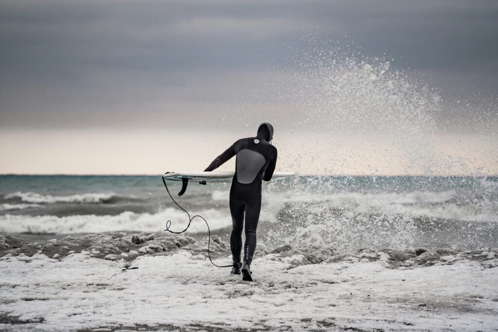 Chilly Water - Cold Water Surf Wax 7:56 PMA surfer wearing a hooded Rip Curl wetsuit and booties walks toward a rough, grey ocean, carrying a white surfboard with a leash trailing behind. The beach is covered in snow and ice, and a large wave crashes dramatically to the right. The overcast sky and wintry conditions convey the harsh cold of a winter surf session - cold water surf wax