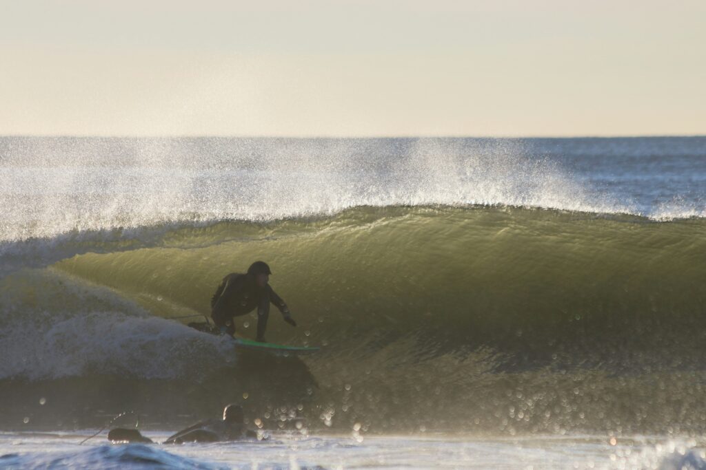 Winter Barrel - Cold Water Surf Wax A surfer in a full black wetsuit crouches low on a teal surfboard inside the barrel of a large, glassy green wave, with white water crashing over the lip. A second surfer is visible in the foreground paddling through the whitewater. The scene is backlit by warm, low-angle sunlight against a pale sky - cold water surf wax