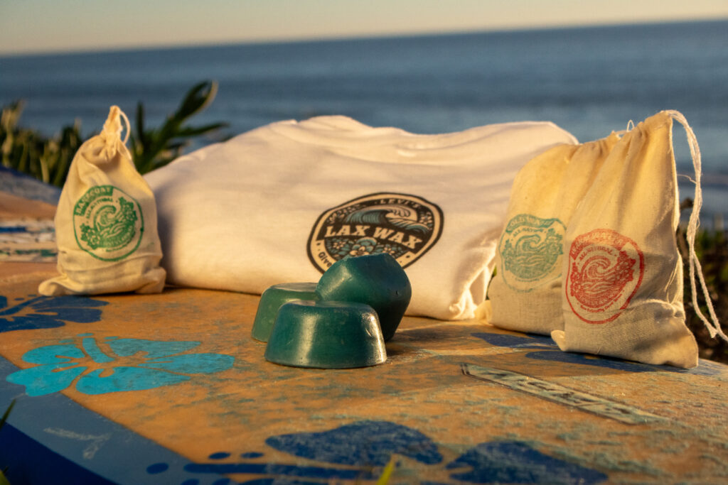 Levi's Lax Wax - Cold Water Surf Wax Two blue cold water surf wax bars rest on a decorated surfboard in the foreground, surrounded by small natural canvas drawstring bags stamped with colorful circular Lax Wax logos in green, blue, and red. A folded white Lax Wax t-shirt sits in the background. The ocean is softly blurred behind the scene, lit by warm golden-hour light.
